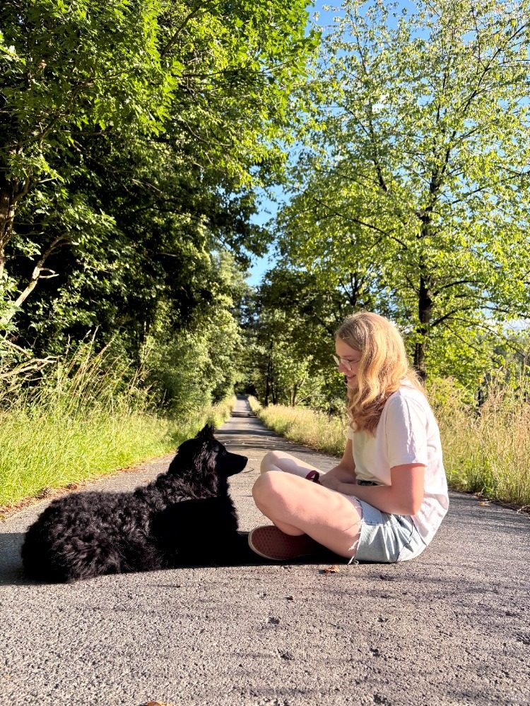 Klara sitzt im Schneidersitz auf einem Waldweg, Pippa liegt ruhig neben ihr